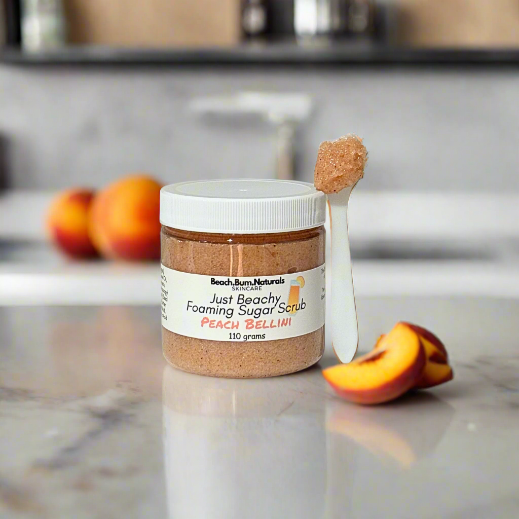Jar of Beach Bum Naturals peach bellini sugar scrub on a kitchen counter with peaches in the background.